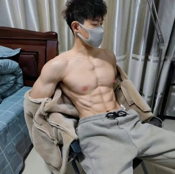 Ảnh hot boy múi có một người đàn ông không mặc áo ngồi trên giường đeo mặt nạ