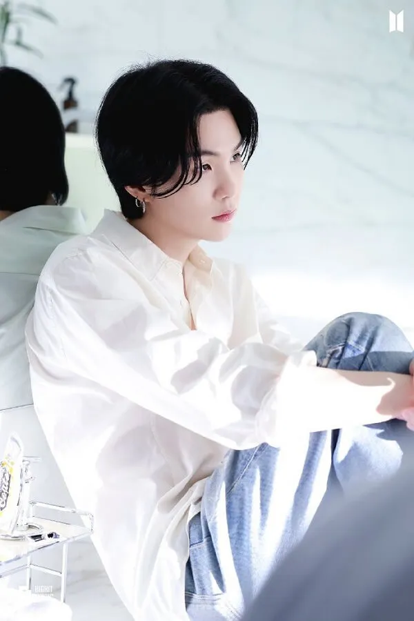 Ảnh suga cute có một người đàn ông đang ngồi trên giường với hai chân của anh ta vượt qua