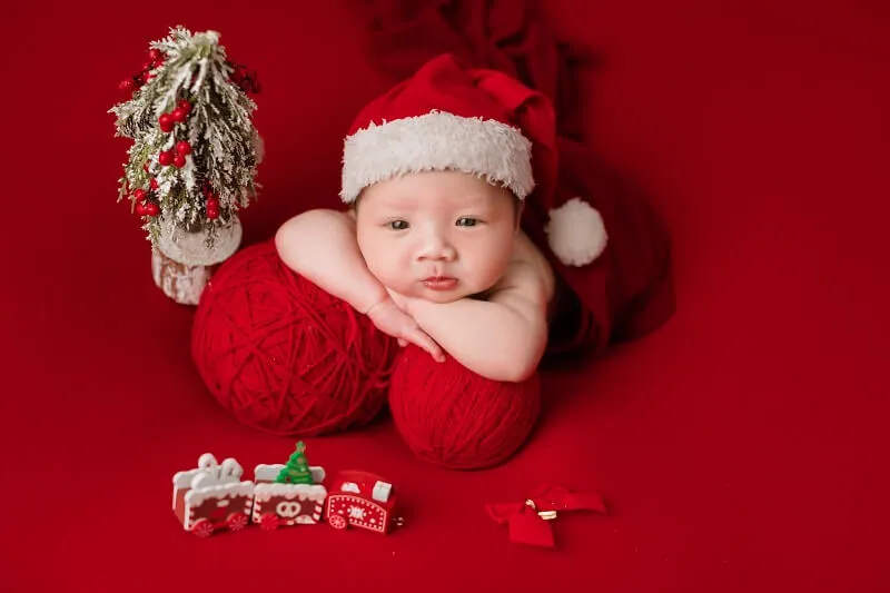 Concept chụp ảnh tết cho be có một em bé đội mũ ông già noel nằm trên quả bóng sợi