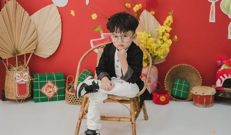 Concept chụp ảnh tết cho bé sơ sinh cậu ngồi trên ghế trước bức tường màu đỏ mặc áo khoác đen và quần trắng