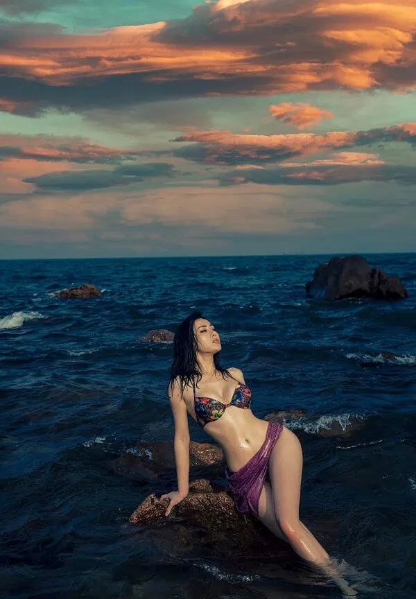 Diễn viên phương oanh người phụ nữ gợi cảm trong bộ bikini màu tím ngồi trên một tảng đá đại dương