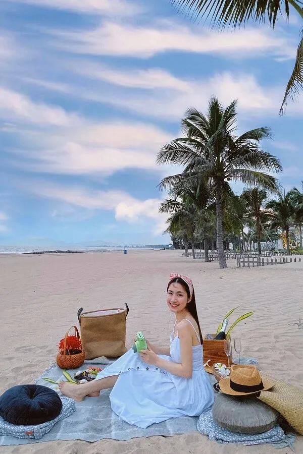Ảnh sex hoà minzy người phụ nữ tóc vàng mặc váy trắng ngồi trên chăn bãi biển với một giỏ đi dã ngoại