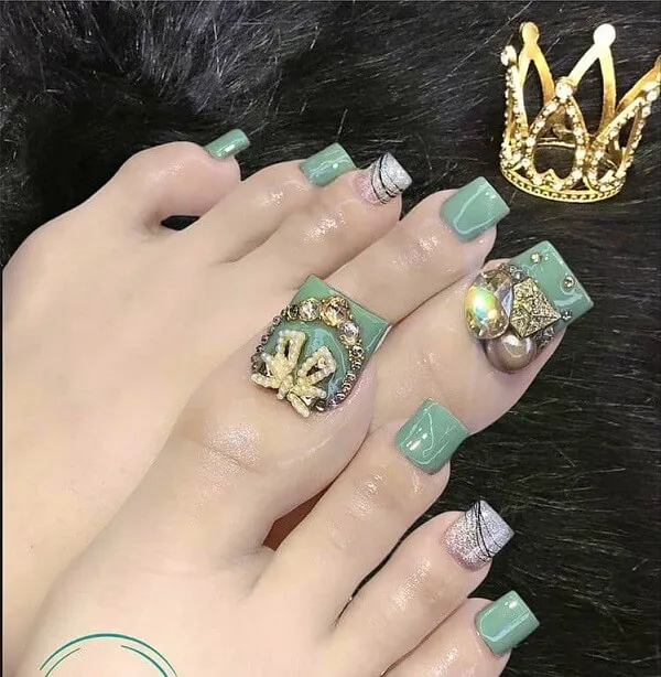 Nail chân đẹp đây là hình ảnh của một người phụ nữ với sơn móng tay màu xanh lá cây và chiếc nhẫn vương miện